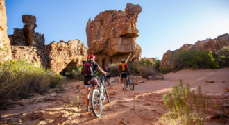 mage showcasing a cyclist traversing the rugged trails of the Cederberg region in South Africa. The cyclist, equipped with a helmet and gear, is captured in motion, illustrating the exhilaration of the ride. The backdrop features dramatic mountain ranges, unique rock formations, and vibrant flora native to the Cederberg area, all bathed in the golden glow of the setting sun. This picture embodies the adventurous spirit of Map2Africa's cycle tours in the captivating Cederberg landscape