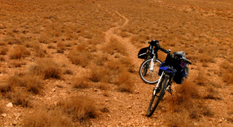 Photograph highlighting a cyclist journeying through the expansive landscapes of the Karoo in South Africa. The cyclist, outfitted with full safety gear, is traveling along a rugged track that meanders through the vast semi-desert terrain. The sparse vegetation, arid plains, and dramatic cloud formations paint a striking picture of the Karoo's stark beauty. This image encapsulates the sense of solitude, resilience, and raw, untouched nature that Map2Africa's cycling holidays in the Karoo region offer
