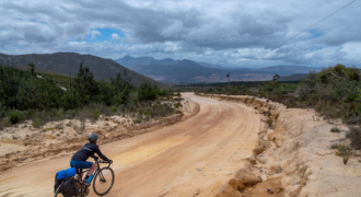 Photograph illustrating a cyclist navigating the picturesque paths of the Overberg region in South Africa. The cyclist, fully equipped with safety gear, is pedaling along a quiet country road flanked by verdant farmland. In the background, majestic mountains stretch towards a clear blue sky, adding a sense of scale and grandeur to the scene. The image captures the serene beauty and diverse landscapes encountered on Map2Africa's cycling tours in the enchanting Overberg region